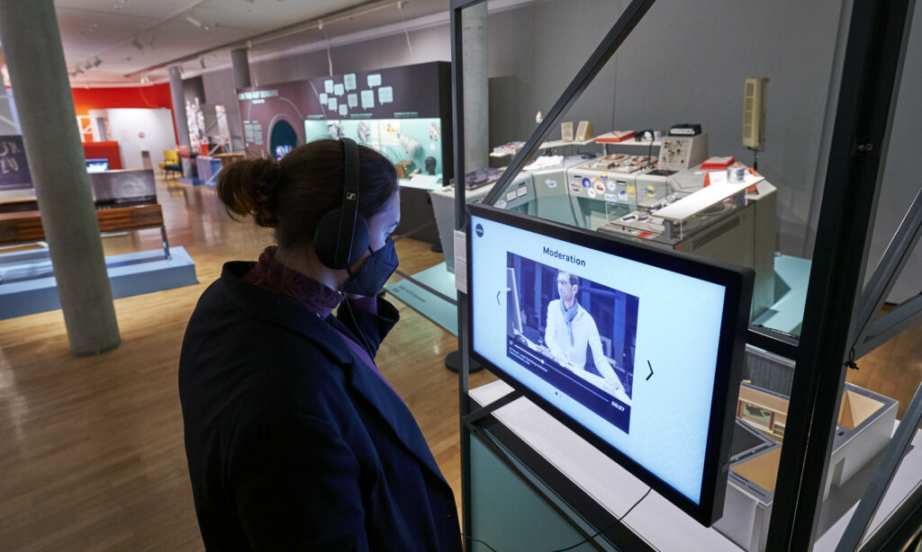 Eine Frau hat Kopfhörer auf und blickt auf eine Medienstation der Ausstellung. Im Hintergrund ist ein Sendeplatz zu sehen.