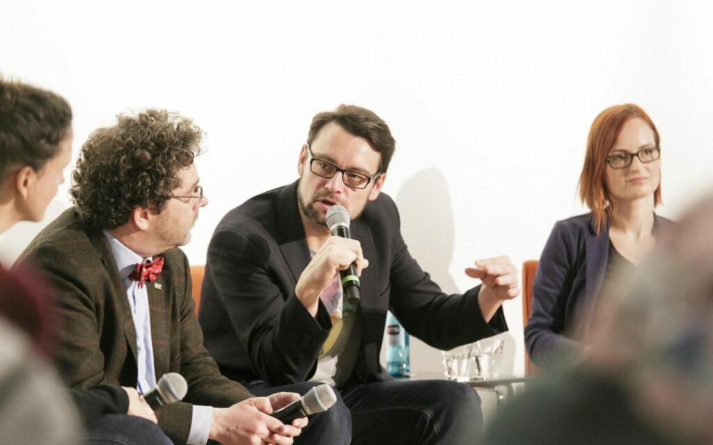 Vier Personen sitzen in einer Diskussionsrunde. Einer führt ein Mikrofon an seinen Mund.