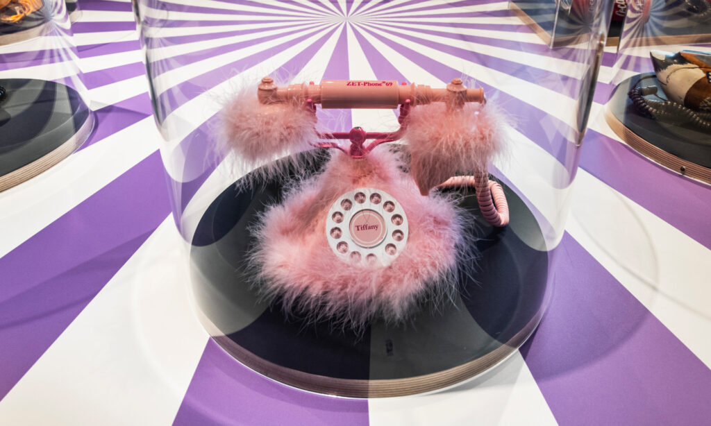 Ein rosa Telefon mit Plüsch steht in einer gläsernen runden Vitrine auf einem lila-weiß gestreiften Tisch.