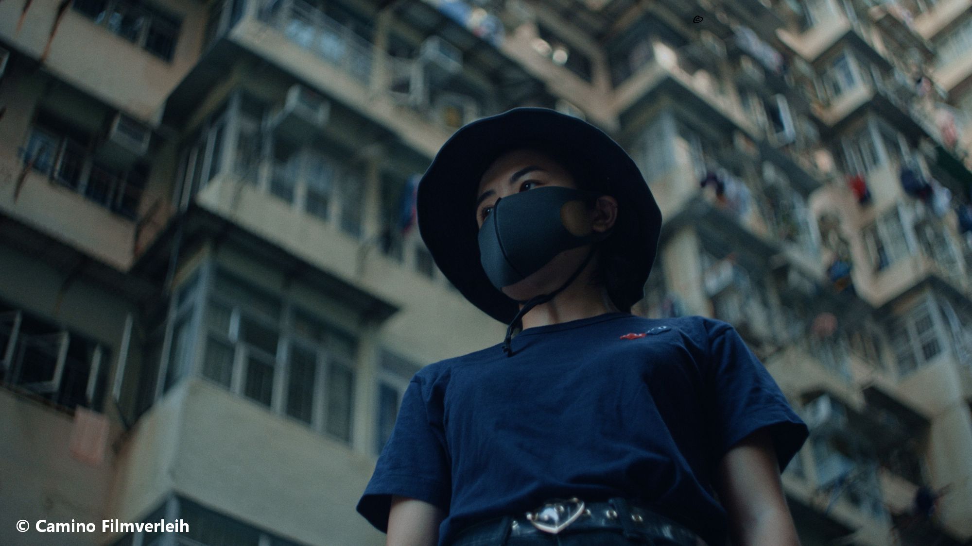 Junge Frau mit blauem T-Shirt und Mund-Nasen-Maske und Hut vor Hochhäusern aus Froschperspetive