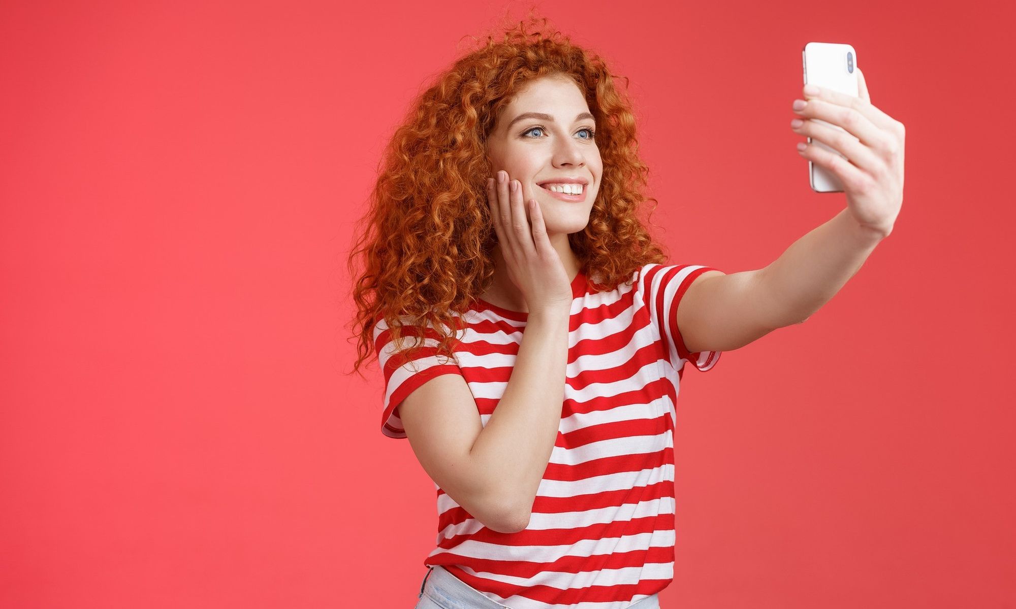 Eine Frau mit langen roten Locken und rot-weiß gestreiftem T-Shirt steht vor rotem Hintergrund und macht ein Selfie von sich