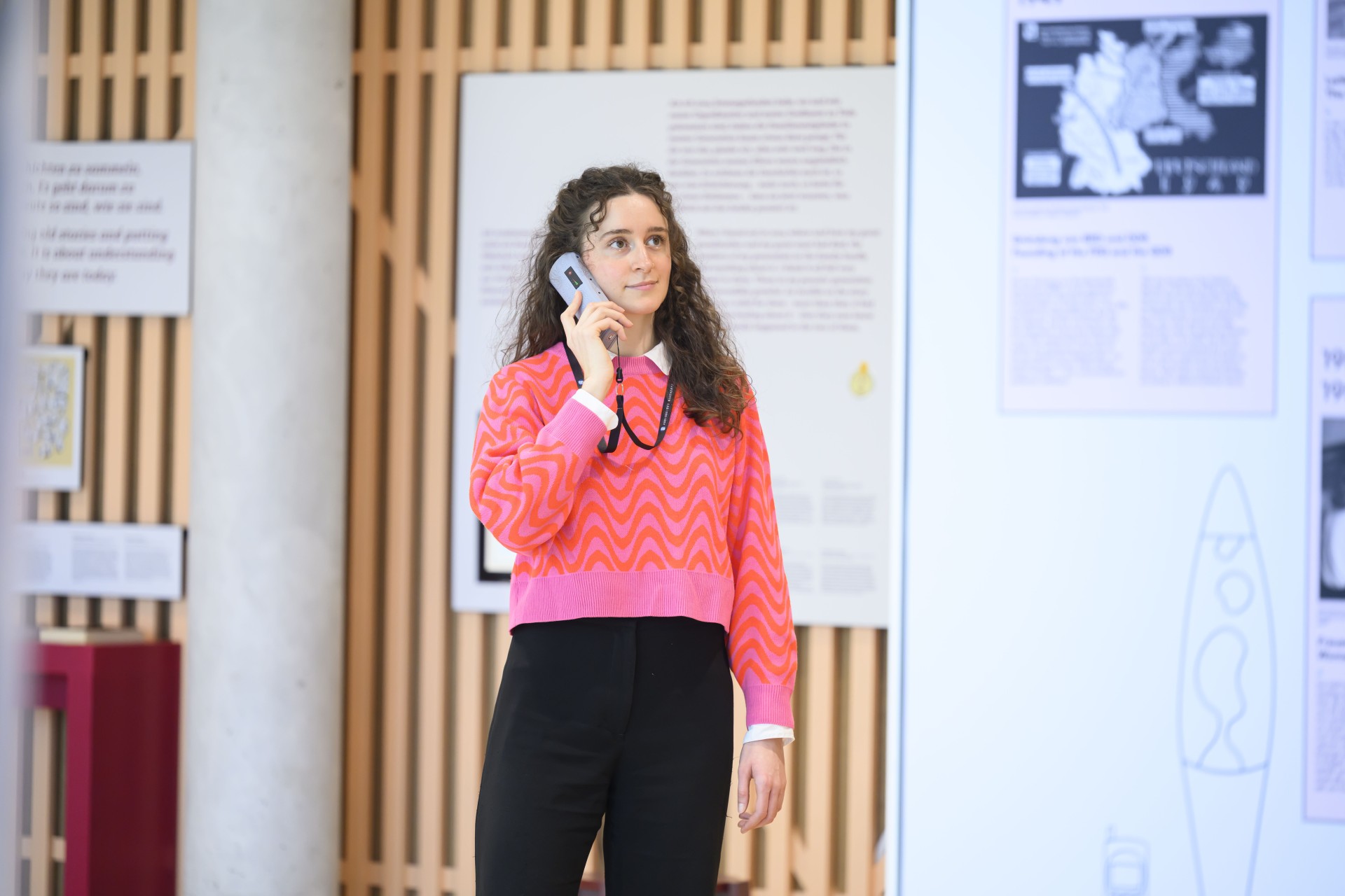 Eine junge Frau mit lockigen, schulterlangen Haaren steht in einem Ausstellungsraum und hält einen Audioguide ans Ohr. Im Hintergrund sind Infotafeln mit Texten und Karten sowie eine Holzlamellenwand zu sehen.