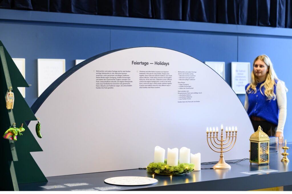 Ausstellungselement in Form eines großen Tisches mit Text und mehreren festlichen Gegenständen wie Kerzenleuchter, Adventskranz, Weihnachtsbaum und Laterne, im Hintergrund eine Frau.