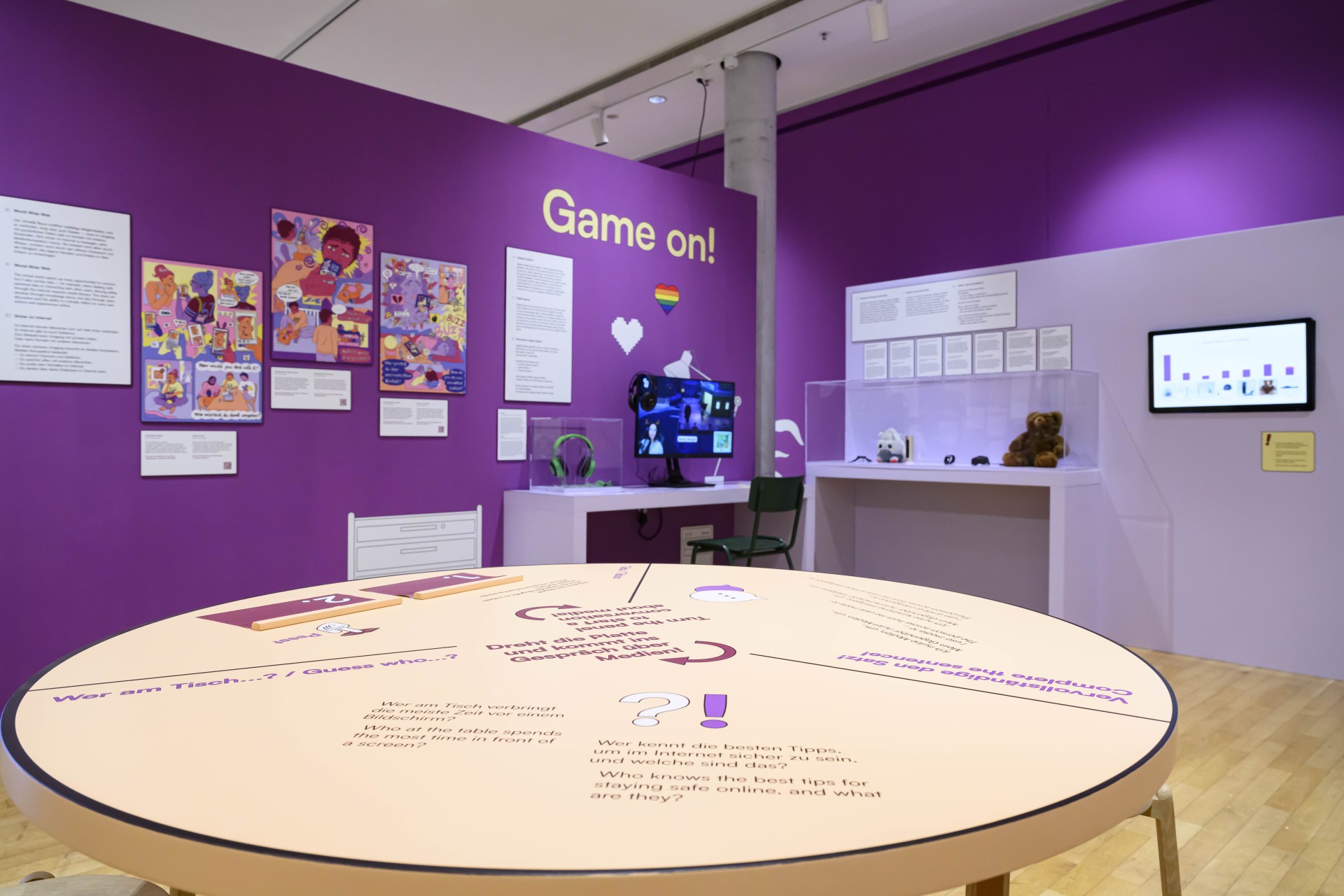 Ein Ausstellungsraum mit lila Wänden zeigt mehrere Informationstafeln, Illustrationen und einen Bildschirm mit Spiele-Controller auf einem Tisch.