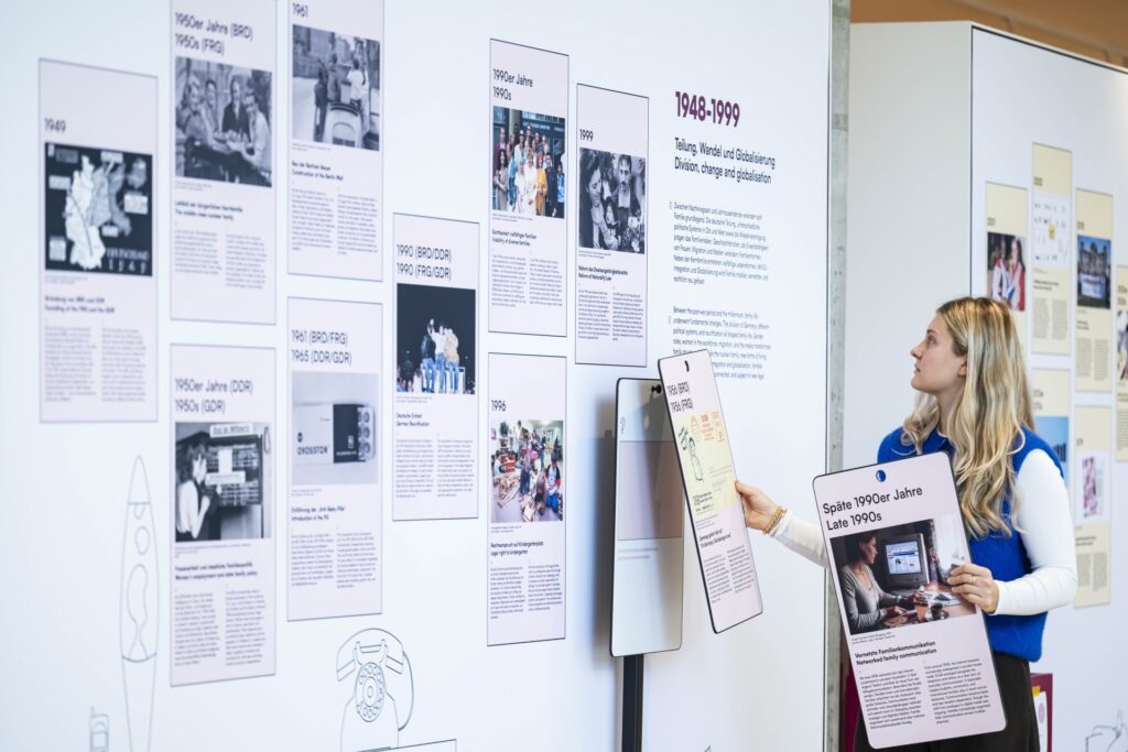 Eine Frau mit langen, blonden Haaren steht seitlich vor einer großen Ausstellungswand. Die Wand ist mit zahlreichen Text- und Bildtafeln bestückt, die chronologisch angeordnet sind. Sie zeigt historische Fotografien, Dokumente und erklärende Texte.