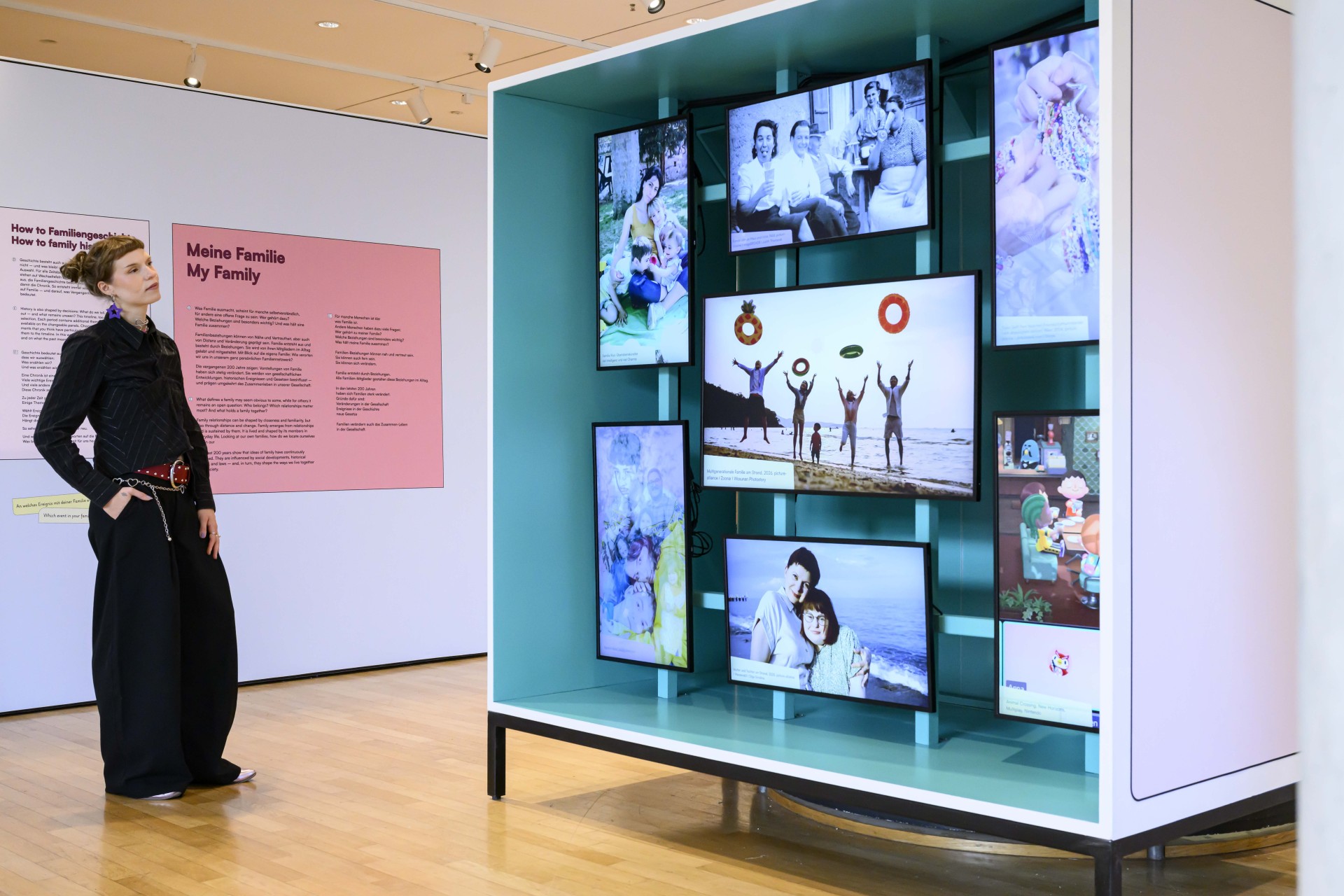 Eine Person steht in einem Ausstellungsraum vor einer großen, türkisfarbenen Vitrine. In der Vitrine sind mehrere Bildschirme in unterschiedlichen Formaten angeordnet, die verschiedene Familienfotos zeigen.