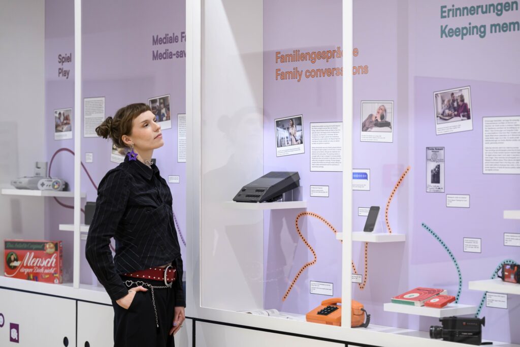 Eine Frau mit kurzem, braunem Haar und dunkler Kleidung steht seitlich vor einer Ausstellungswand mit mehreren Vitrinen. Die Vitrinen sind in Pastellfarben gehalten und enthalten verschiedene Mediengeräte, darunter ein altes Telefon, eine Spielkonsole und ein Diktiergerät.
