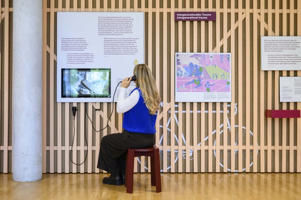 Eine Person mit langen, offenen Haaren sitzt auf einem Holzstuhl vor einer Ausstellungswand, die an einen Kellerverschlag erinnert. Sie hält einen Telefonhörer ans Ohr, während sie auf einen Monitor blickt.