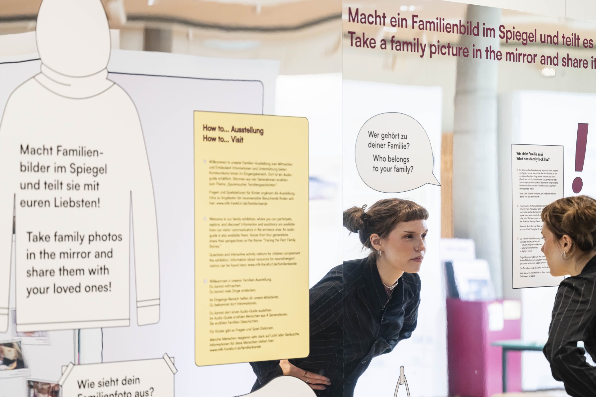 Eine junge Frau mit kurzem, braunem Haar steht vor einem großen Spiegel und betrachtet ihr Spiegelbild. Im Vordergrund befinden sich mehrere aufgestellte Tafeln mit Texten in Deutsch und Englisch. Die Tafeln sind in hellen Farben gehalten und enthalten Anweisungen sowie Fragen zur Familienzugehörigkeit.