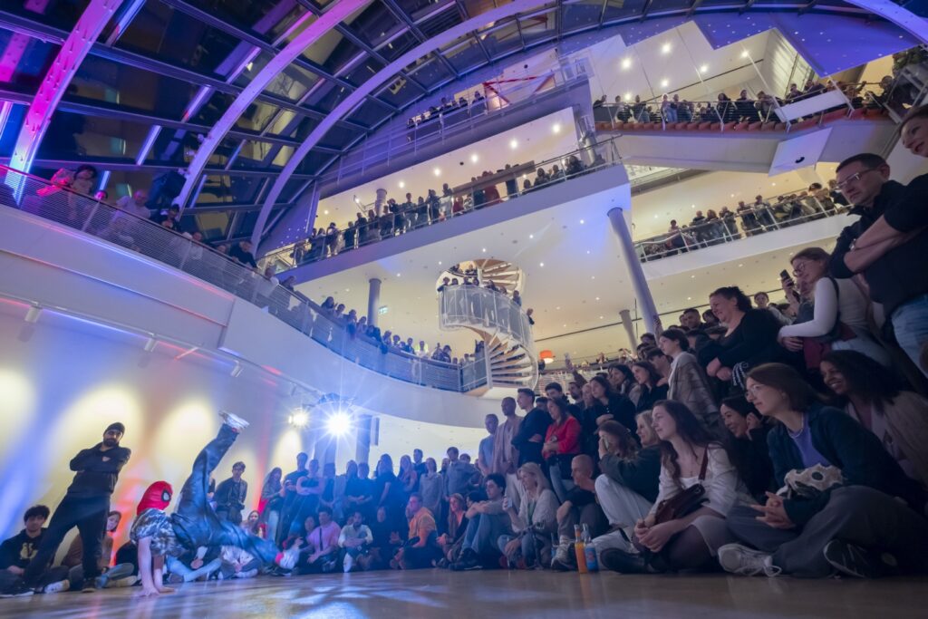 Blick vom Forum des Museums aus einer bodennahen Perspektive in das Museum, sodass die Architektur des Hauses mit Treppen, Wendeltreppen und Glasdach zu erkennen ist. Das Forum, die Gänge und Treppen sind voller Menschen. Links ist eine Breakdance-Gruppe zu sehen. Das Museum ist blau-rosa ausgeleuchtet.