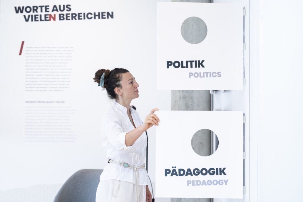 Frau in weißer Bluse steht vor zwei weißen Schildern mit den Aufschriften 'POLITIK' und 'PÄDAGOGIK' in einer Ausstellung.