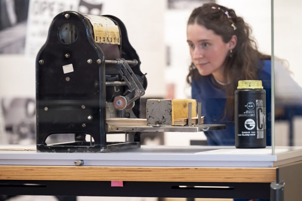 Frau betrachtet hinter Glas ein historisches mechanisches Gerät mit Papierrolle und Farbband in einer Ausstellung.
