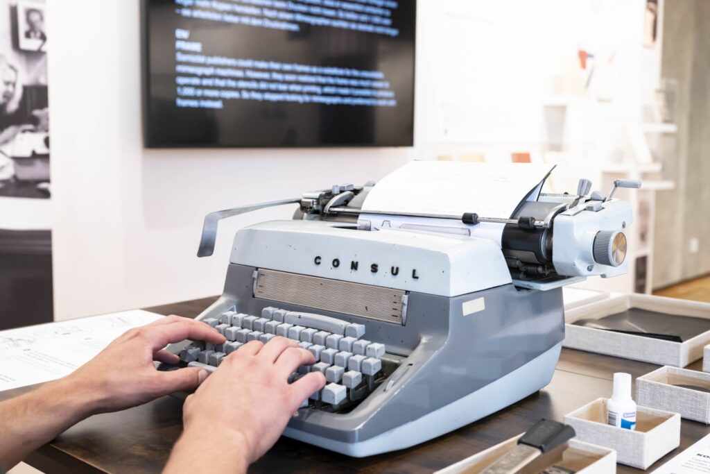 Hände tippen auf einer grauen Schreibmaschine der Marke Consul auf einem Tisch in einem Ausstellungsraum.