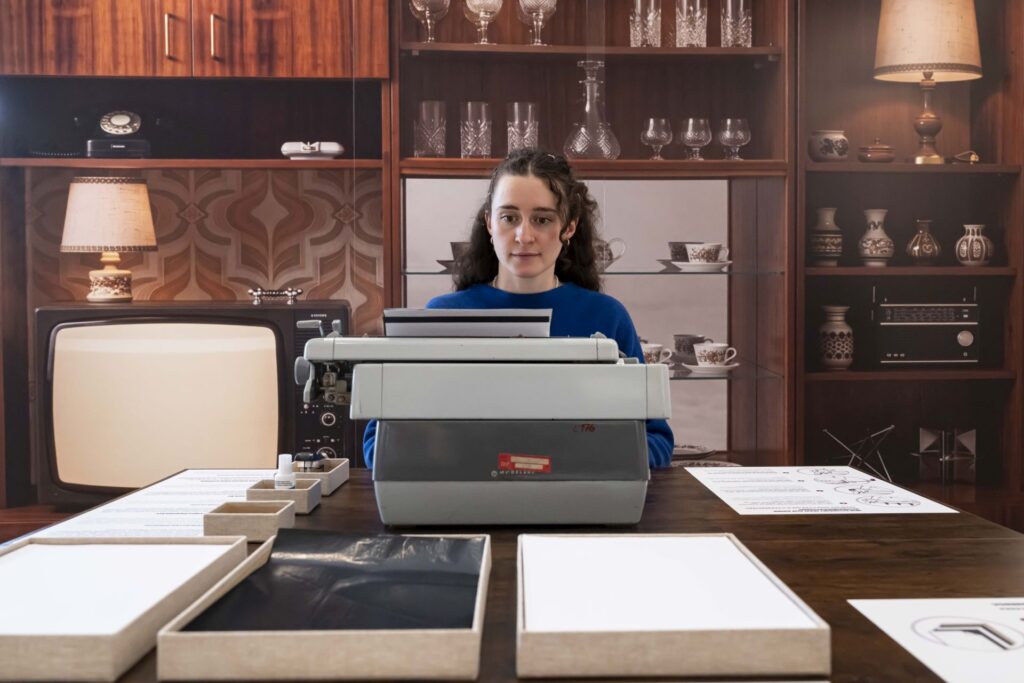 Frau mit dunklen Haaren sitzt an einem Tisch vor einer Schreibmaschine in einem Raum mit Retro-Möbeln und -Dekorationen