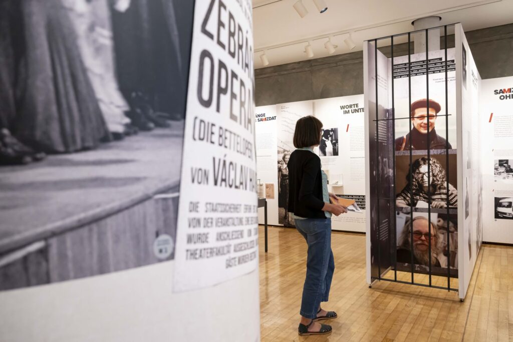 Person steht in einer Ausstellung vor einer Gitterstruktur mit Porträts und Texten an den Wänden und hält ein kleines Heft in der Hand.