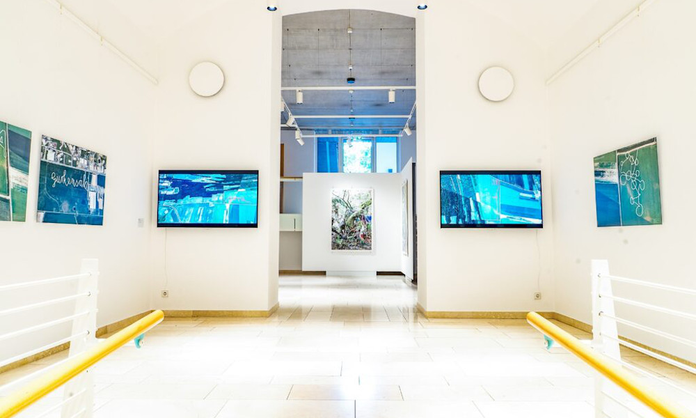 Ausstellungsansicht der Kunsträume aus der Eingangsperspektive. An den Wänden hängen Bilder und zwei Bildschirme