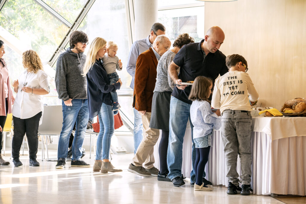 Menschen unterschiedlichen Alters und Geschlechts stehen in einer hellen, modernen Umgebung in einer Schlange vor einem Buffet mit Brot und Bananen