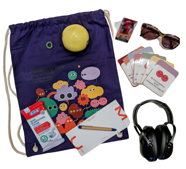 Verschiedene Utensilien neben einem lilafarbenen, mit Monster-Illustrationen bedruckten Beutel: Darunter Kopfhörer, eine Sonnenbrille, ein Anti-Stress-Ball und kleine Kärtchen.