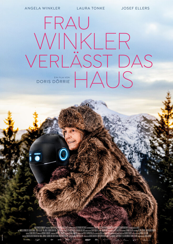 Frau in braunem Fellmantel umarmt einen schwarzen Roboter vor schneebedeckten Bergen mit blauem Himmel