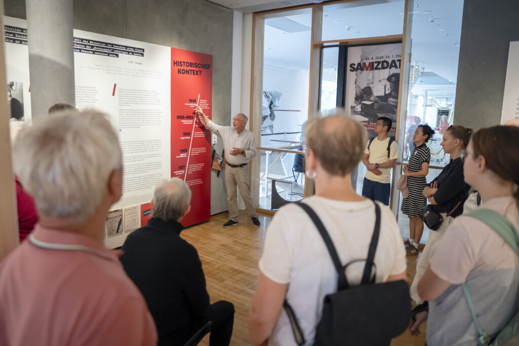 Ein Mann erklärt einer Gruppe von Menschen in einem Ausstellungsraum eine Wand mit Texttafeln und einer roten Zeitleiste.