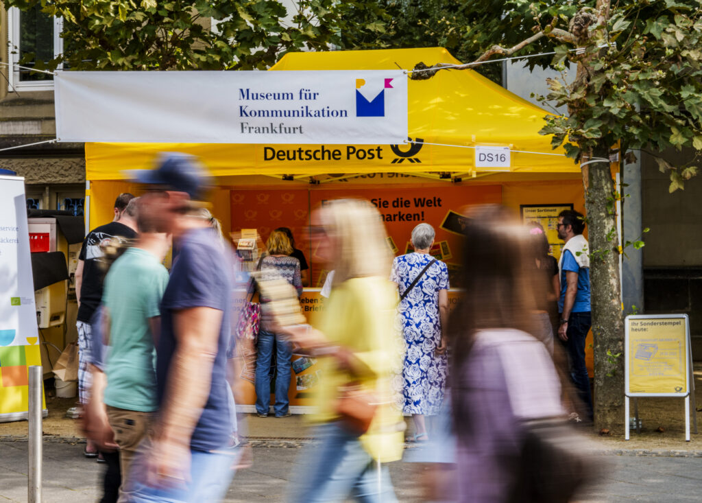 Gelber Pavillon mit Aufschrift 'Museum für Kommunikation Frankfurt' und 'Deutsche Post' vor Baum mit mehreren Menschen in Bewegung im Vordergrund


