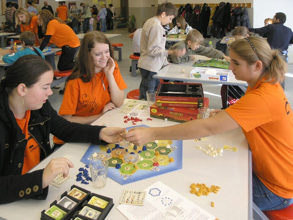 Drei junge Frauen in orangefarbenen T-Shirts sitzen an einem Tisch und spielen das Brettspiel Siedler von Catan mit Spielmaterialien und Karten