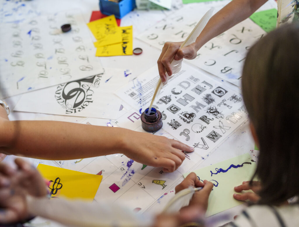 Mehrere Personen sitzen an einem Tisch mit Papieren voller Buchstaben und kalligrafischen Zeichen, eine Person taucht einen Pinsel in ein Tintenfass

