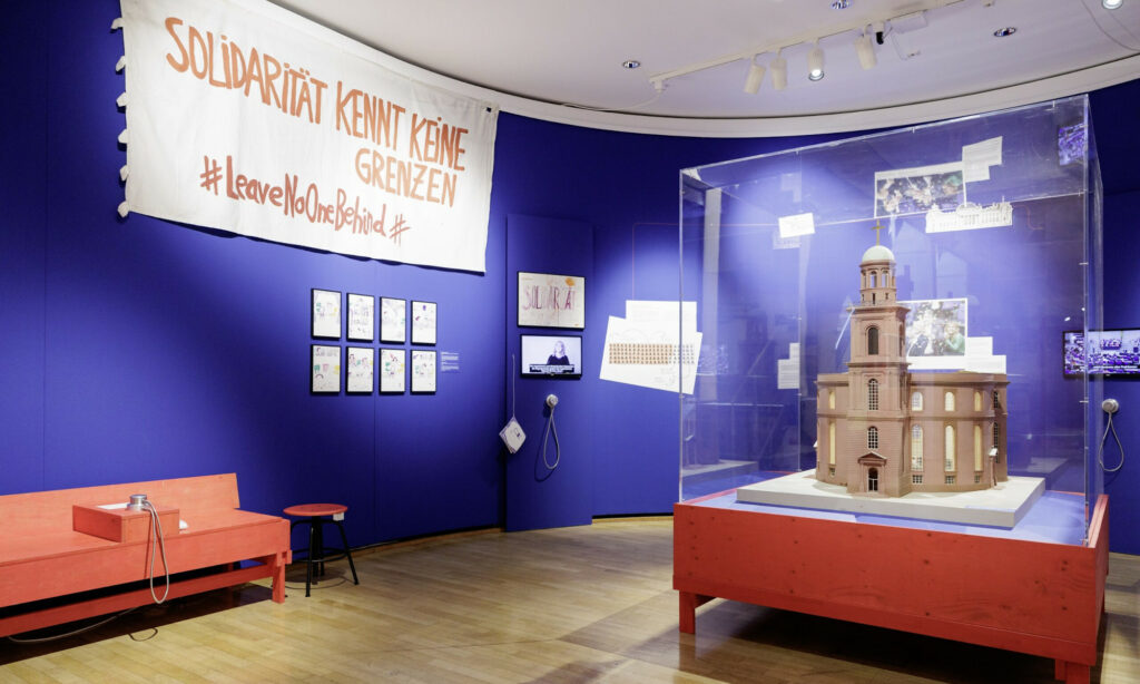 Blick in die Ausstellung "Streit. Eine Annäherung" im Museum für Kommunikation Frankfurt. Zu sehen ist ein Modell der Paulskirche und ein Banner mit dem Text "Solidarität kennt keine Grenzen #LeaveNoOneBehind"