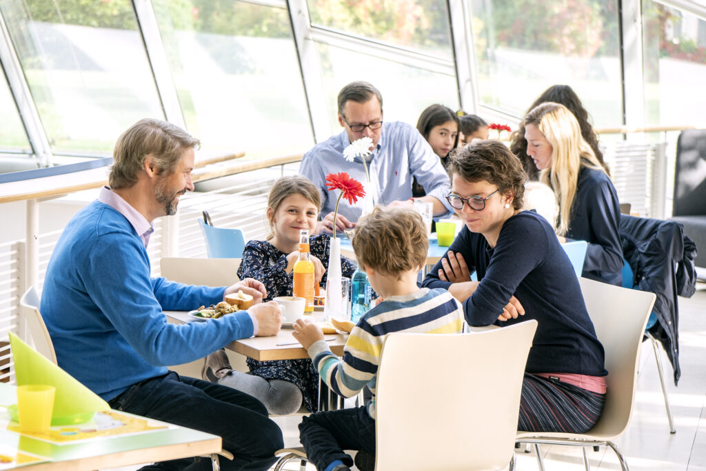 Helle Cafészene mit mehreren Personen an weißen Tischen. Im Vordergrund sitzt eine Familie mit zwei Kindern, die gemeinsam frühstückt. Auf dem Tisch stehen Teller mit Speisen, Gläser mit Saft und eine weiße Vase mit einer roten Blume. Im Hintergrund unterhalten sich weitere Gäste. Große Fenster lassen viel Tageslicht herein und bieten einen Blick ins Grüne.