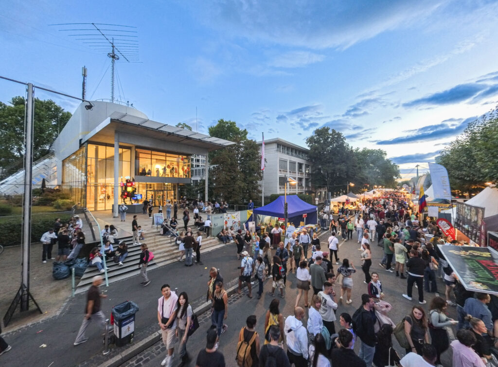 Menschenmenge bei einem Straßenfest vor dem Museum für Kommunkation mit großen Fenstern und Treppen unter blauem Himmel am Abend