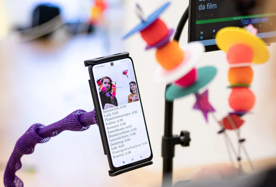 Smartphone in Halterung zeigt Videoanruf mit zwei Personen und Textliste, im Vordergrund bunte, unscharfe Objekte